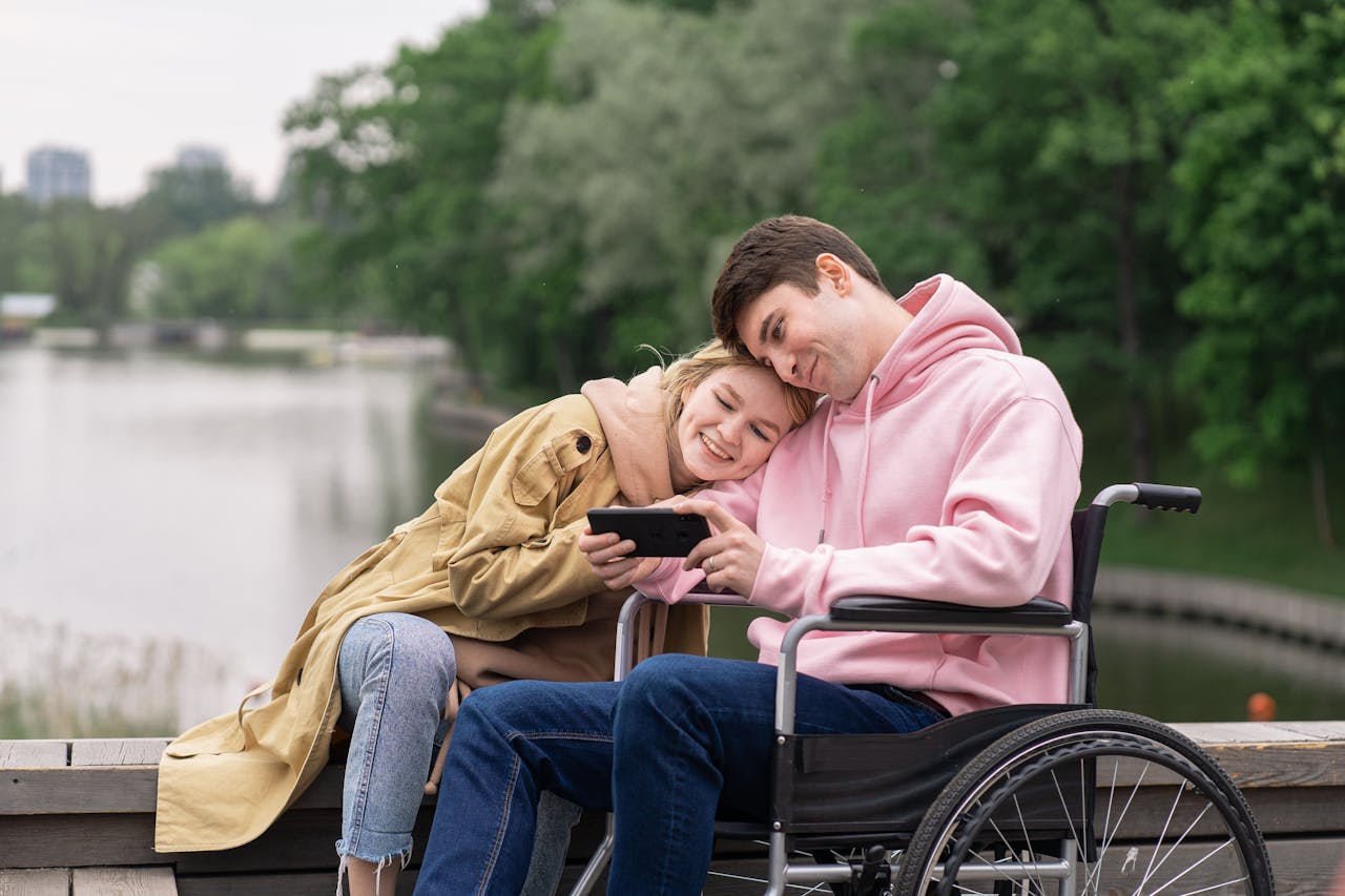 Smiling couple by lakeside enjoying a moment on smartphone, one in a wheelchair.